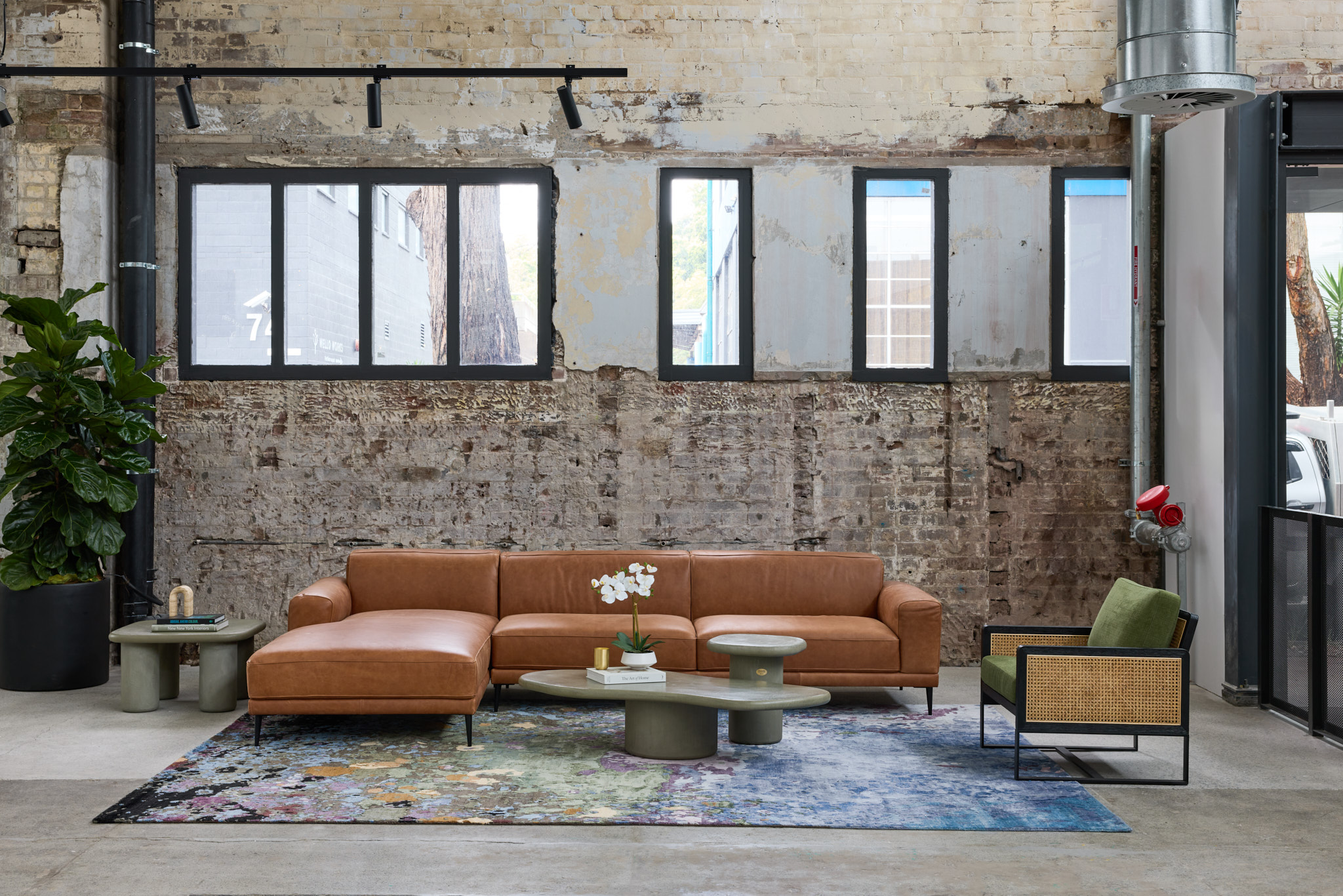 Luma Developments The interior design features a modern living room with concrete flooring, brick walls, large windows, and furnishings including a brown leather sofa, green chair, colorful rug, and a potted plant.