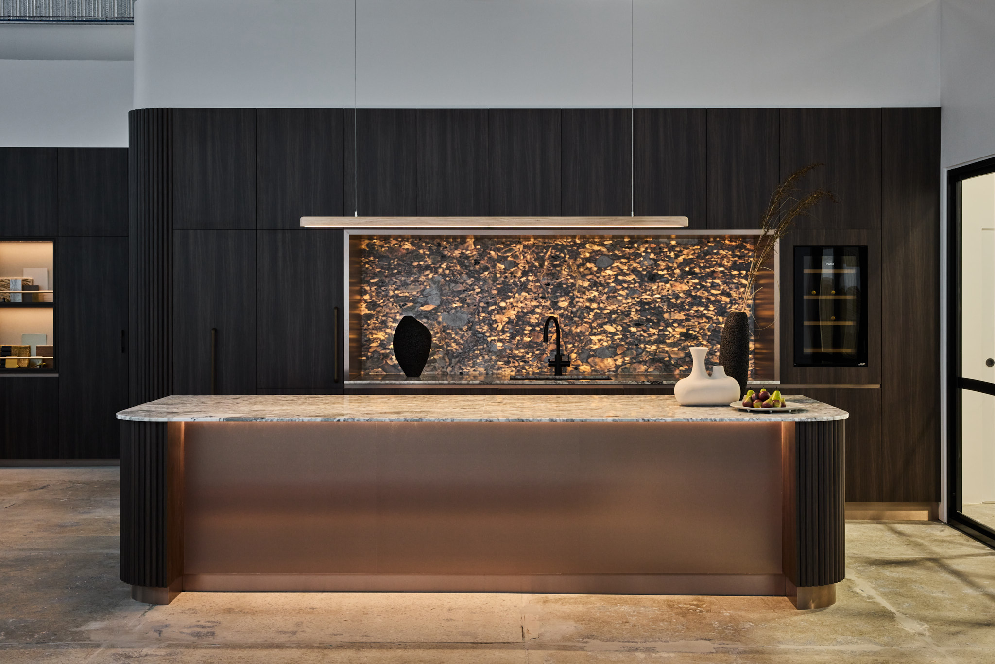 Luma Developments A modern kitchen features a large marble island, dark wood cabinetry, and a decorative backsplash with a hanging light fixture above.